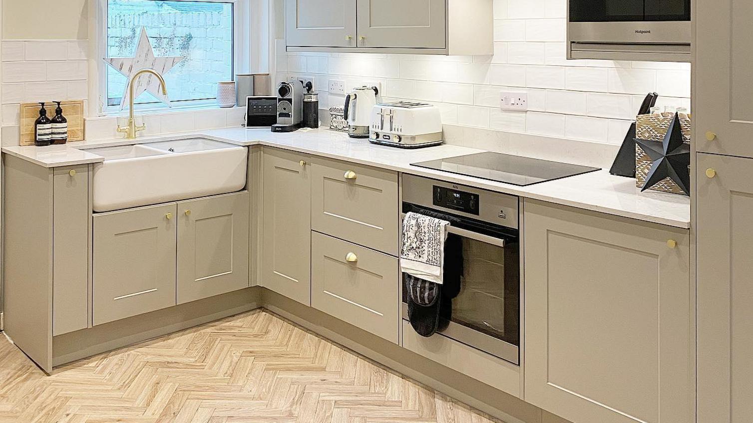 A luxurious pebble kitchen idea featuring shaker doors, white worktops, a Belfast sink, a brass tap, and herringbone flooring.