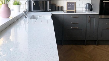 A luxurious dark-grey L-shaped kitchen idea that has white quartz worktops, chevron flooring, and an undermount sink.