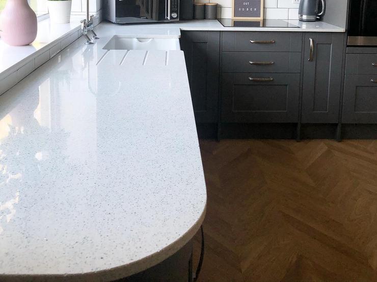 A luxurious dark-grey L-shaped kitchen idea that has white quartz worktops, chevron flooring, and an undermount sink.