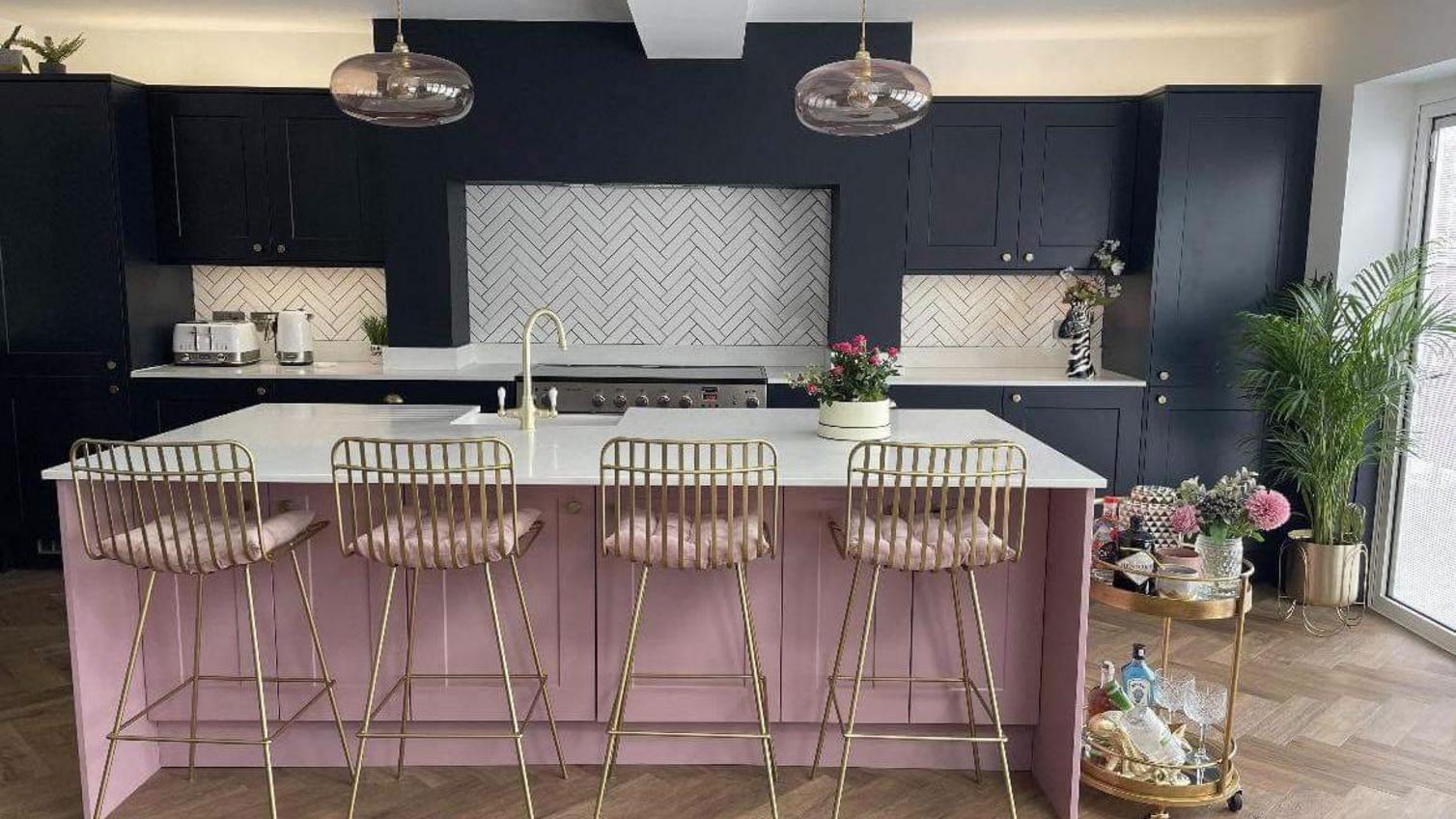 Two tone paintable kitchen design with a pink island, navy units, chevron flooring, gold stools, and white chevron tiles.