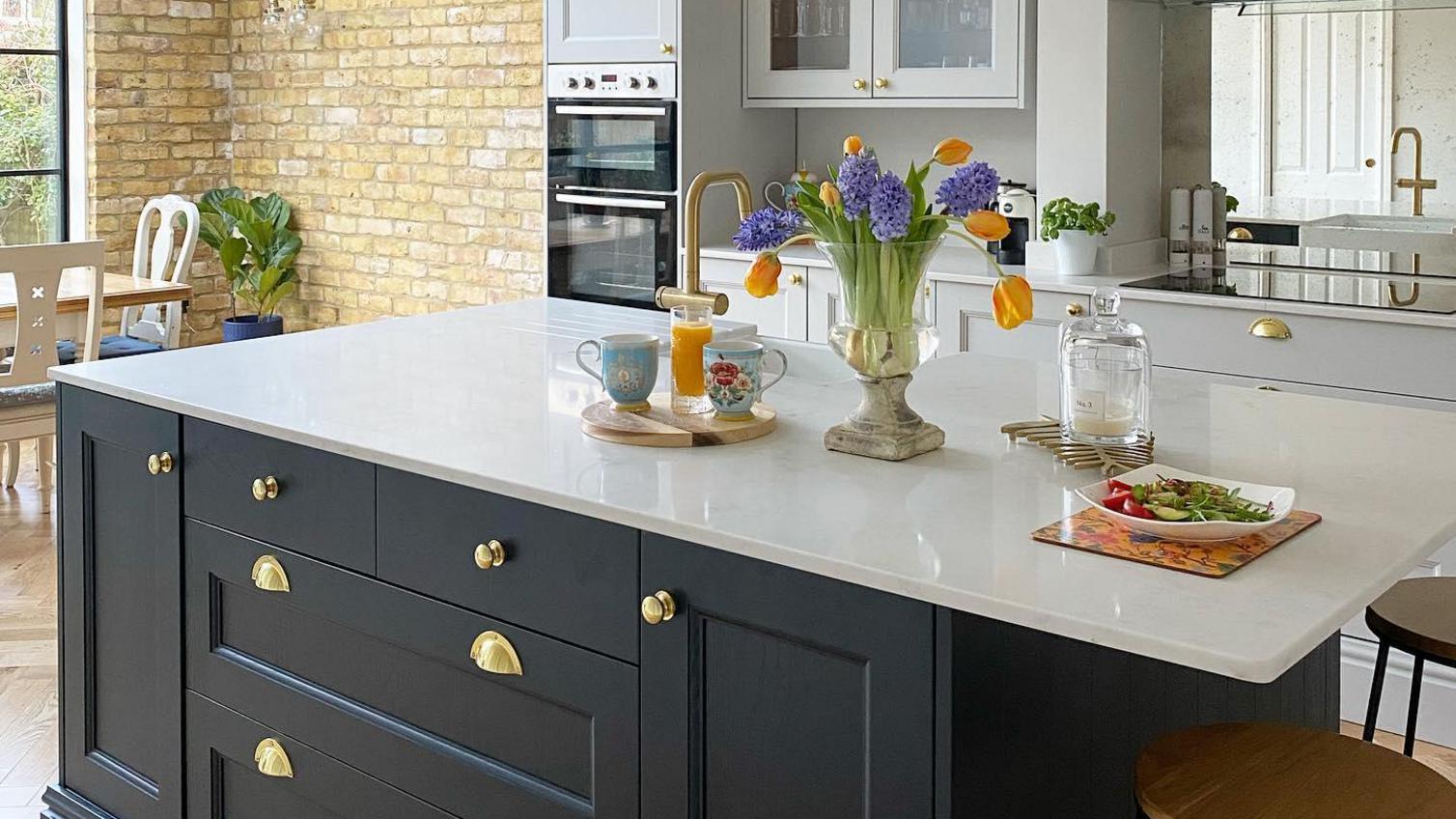 Two-tone kitchen idea with a navy island, dove-grey wall units gold cup handles, a white worktop, and decorative objects.