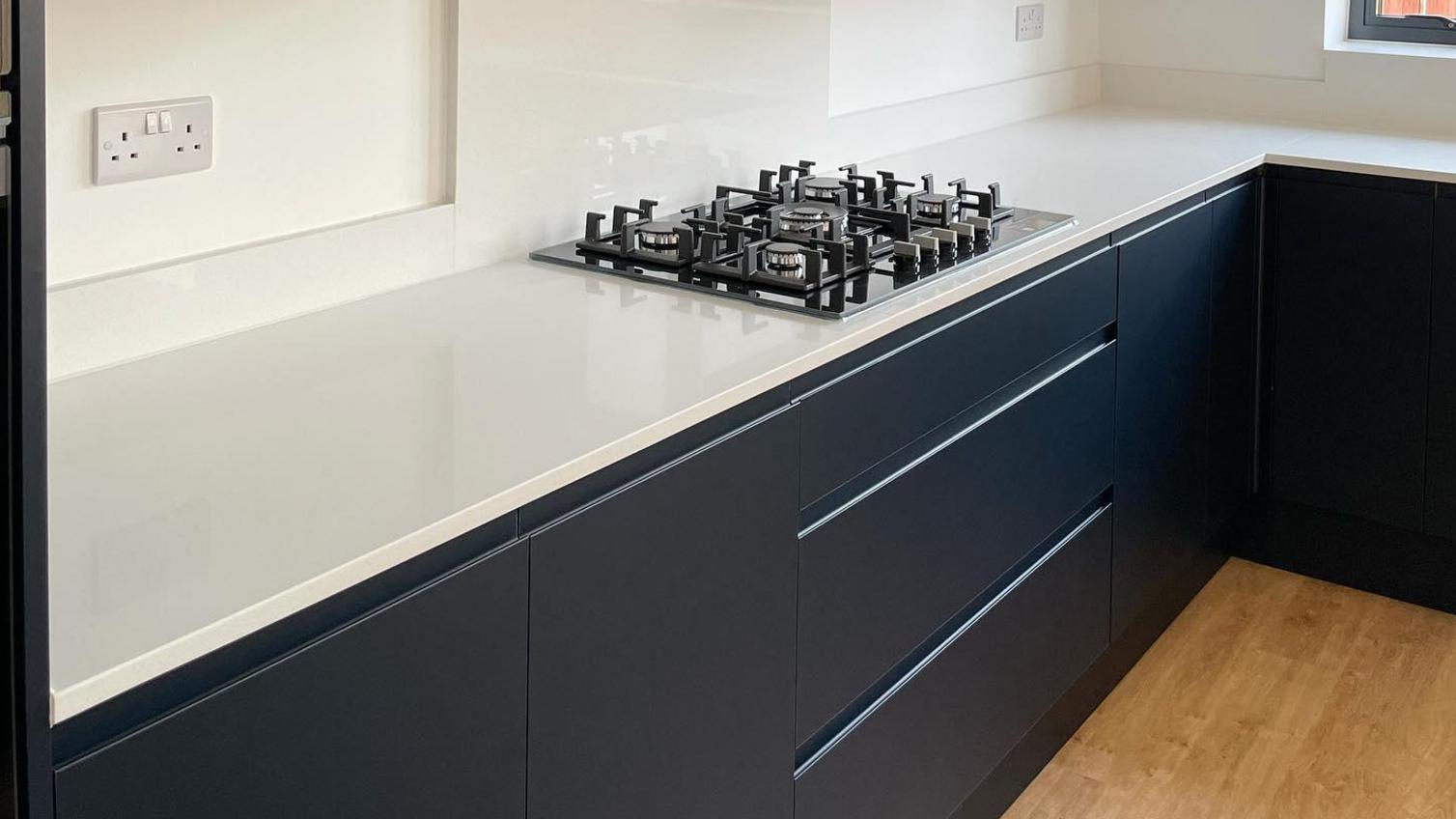 Navy integrated handle kitchen in a u-shaped layout with white worktops, a silver extractor, black hob, and wood floors.