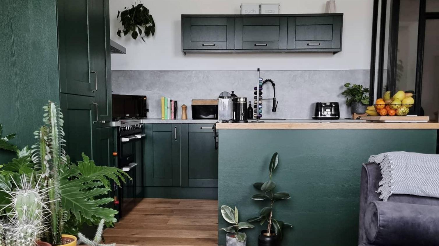 Dark green kitchen island design with brass bar handles, oak worktops, oak flooring, and a living room in shot.