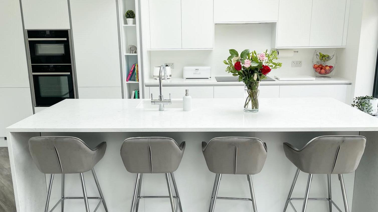 white gloss open plan kitchen with slab doors, white wraparound island worktop and grey stools.