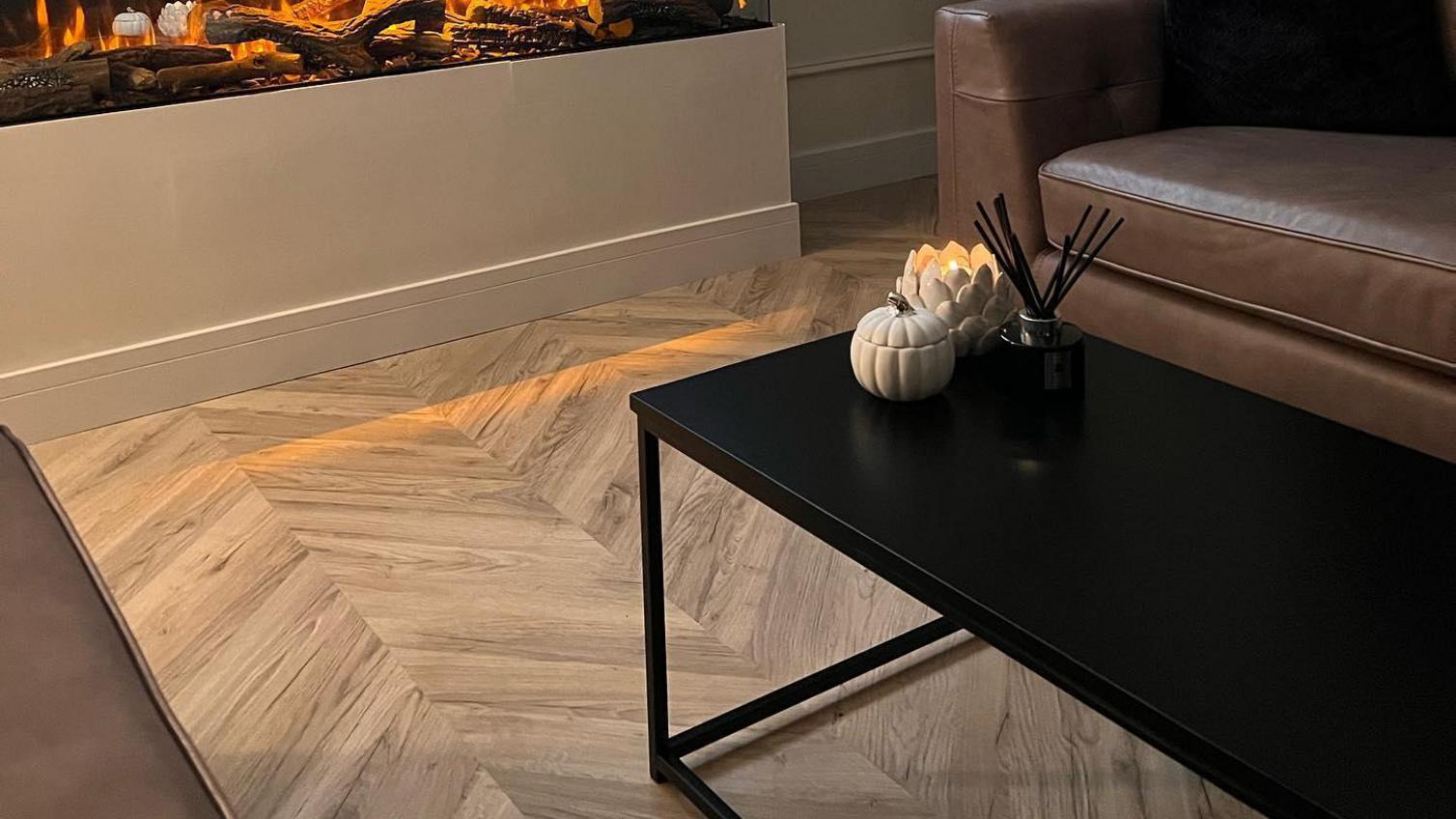 A luxe living room idea using oak chevron flooring. Contains a black coffee table, a brown leather sofa, and white skirting.