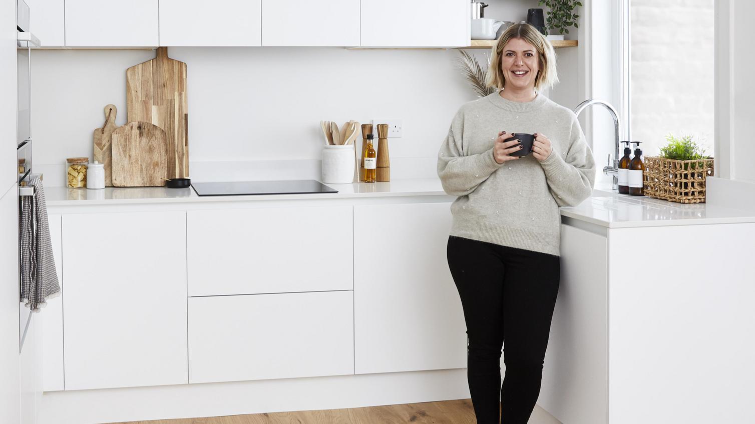 Woman standing in an u-shaped modern white kitchen. Has wooden flooring, white quartz worktops, and tower units.