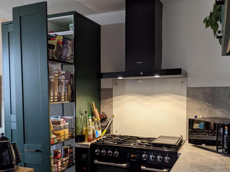 Green shaker kitchen design with black hob and extractor, larder style pull out cupboard, and grey concrete-effect worktops.