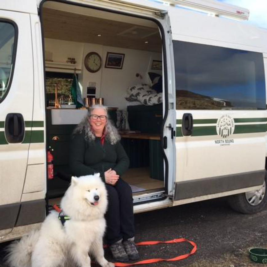 Sharon sat down on the edge of the campervan door, with her fluffy white dog. Showing the interior and side of the campervan.