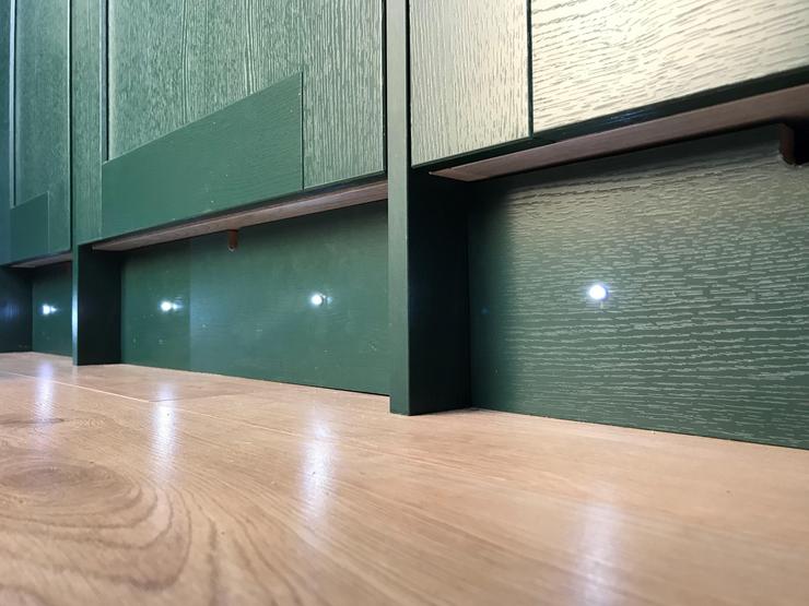 Plinth lights on the bottom of Fairford dark green cupboards in a campervan, showing them lit up next to light oak flooring.