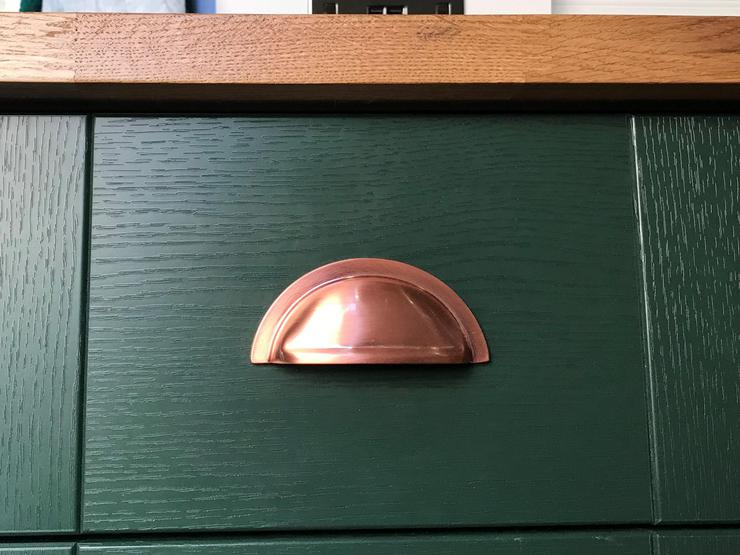 Copper cup handle on a Fairford dark green shaker cupboard, showing copper hinges, wood-effect worktop and white panelling.