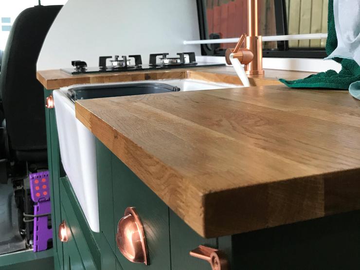Edge of wood-effect laminate worktop with a white Belfast sink, copper tap and handles, and dark green shaker cupboards.