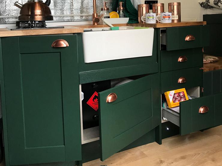 Cooking space in van with dark green shaker doors open showing storage options, such as drawers and under-sink cupboards.