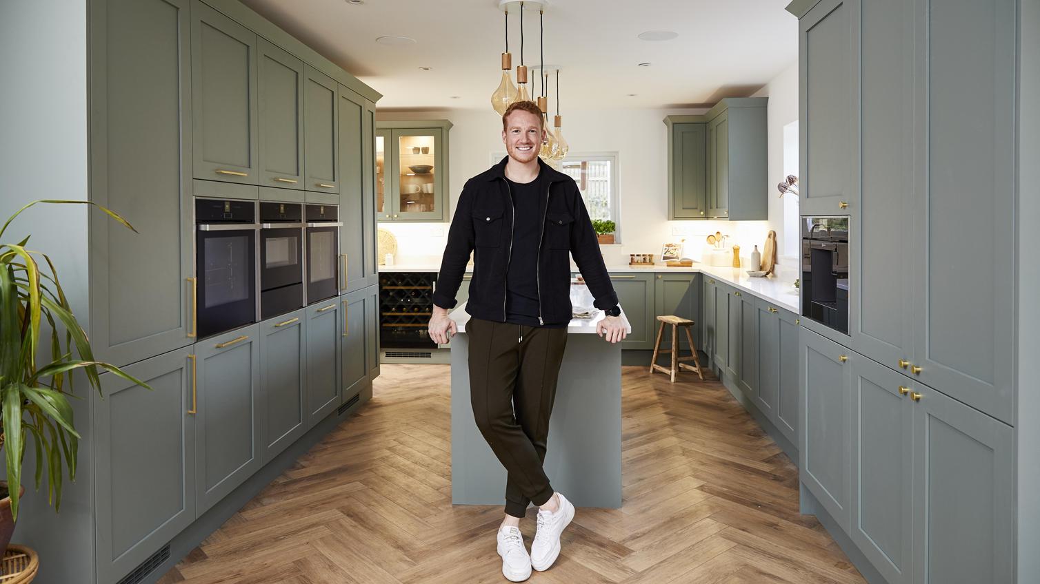 Greg Rutherford stood at the end of a kitchen island, showing sage green shaker cupboards and chevron wood flooring.