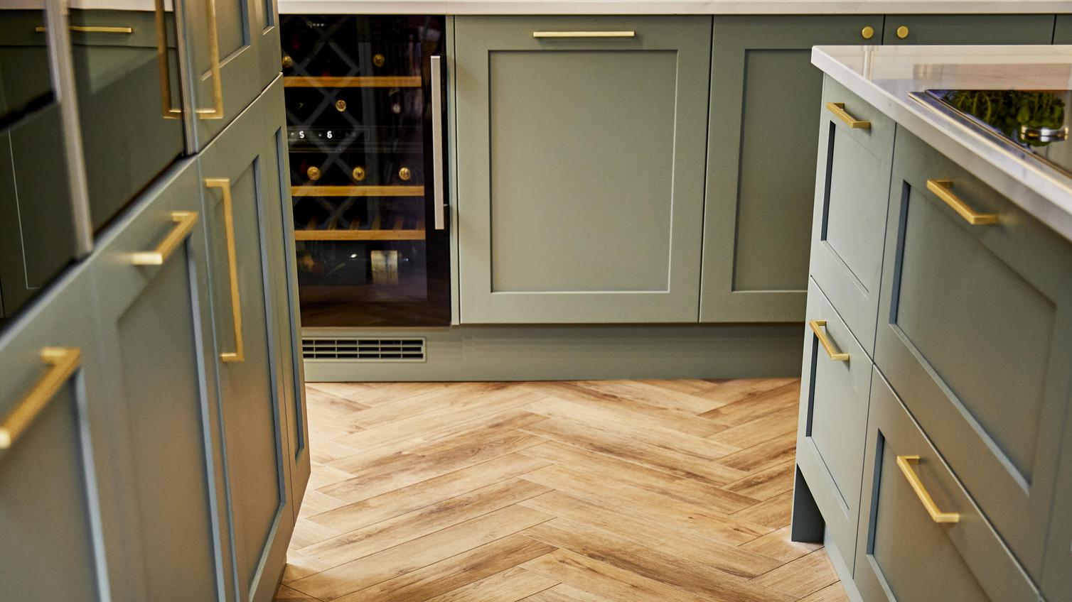 Sage green shaker kitchen design with antique wood chevron flooring, brass bar handles, and a wine fridge.