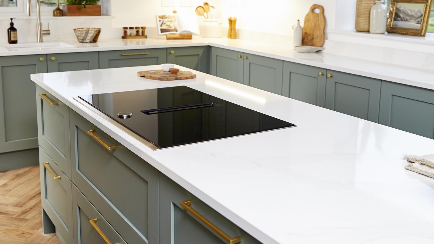 Sage green shaker kitchen in an island layout with brass bar handles, white worktops, and an induction hob.