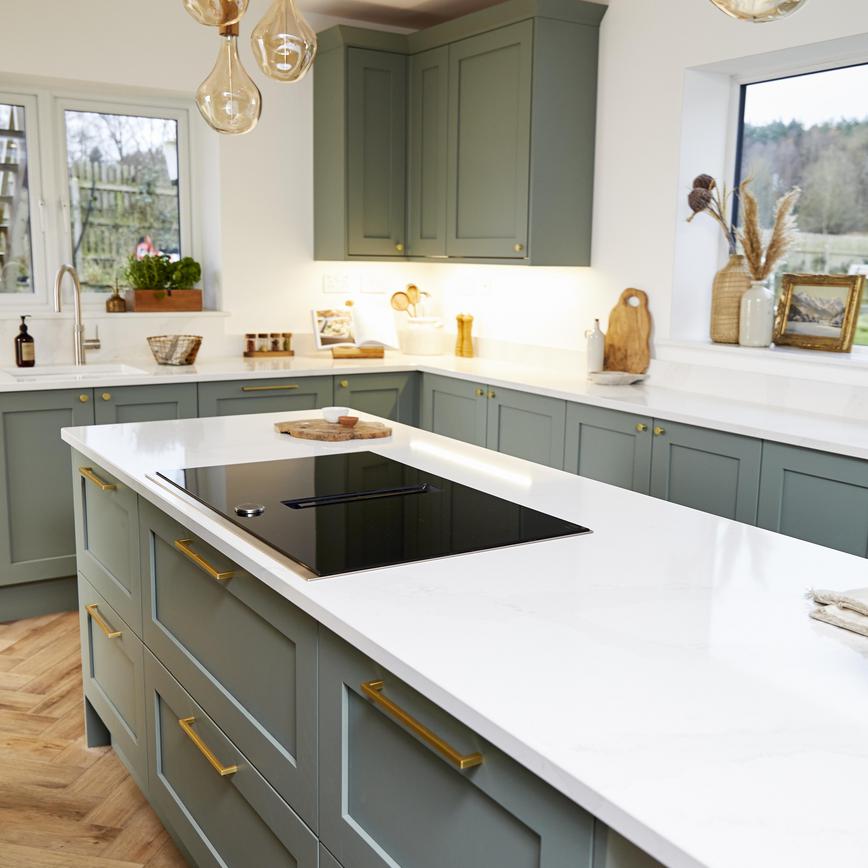 Sage green shaker kitchen in an island layout with brass bar handles, white worktops, and an induction hob.