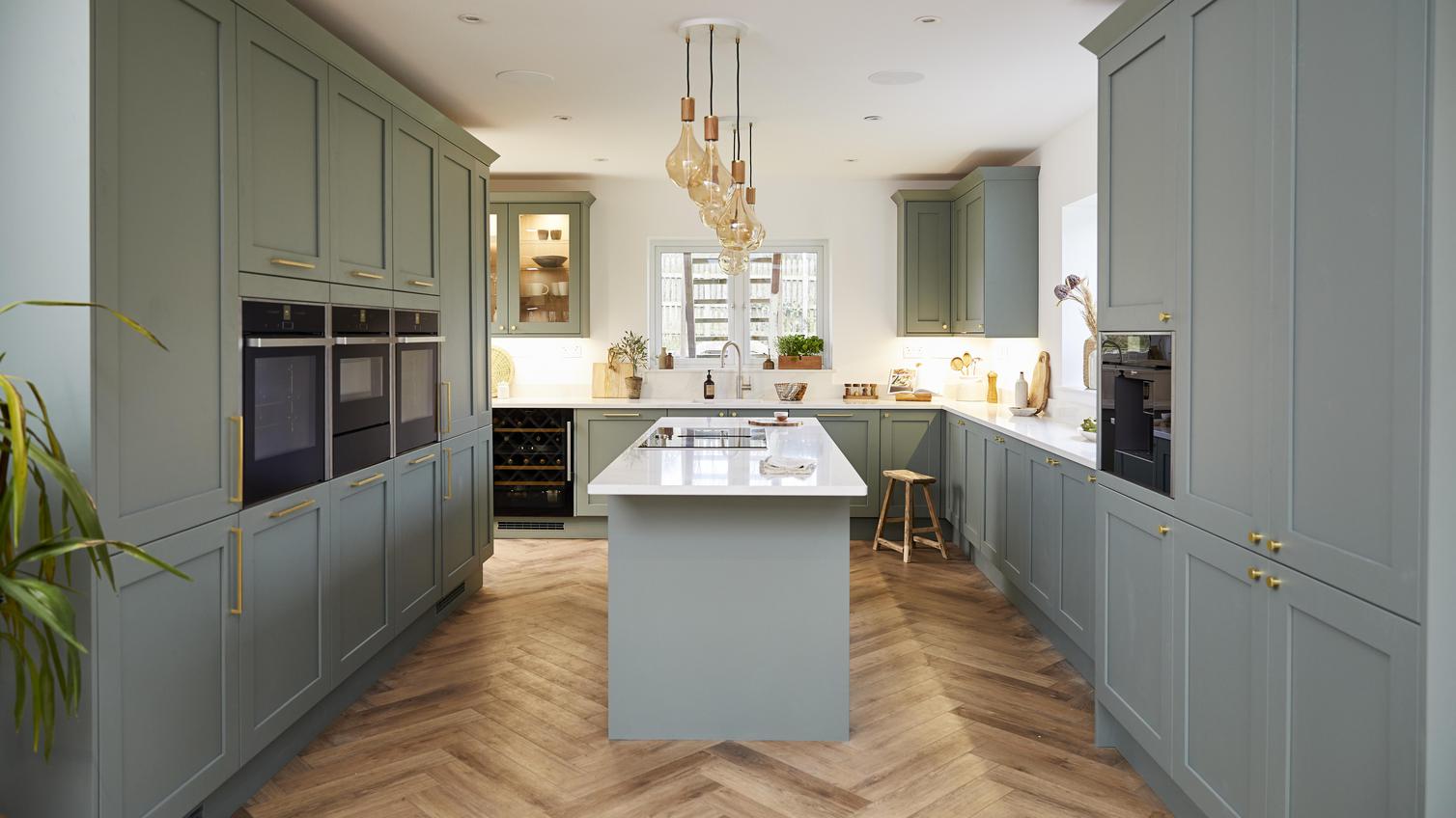 Sage green shaker kitchen in an island layout, with wooden chevron floors, brass handles, white worktops and pendant lights.