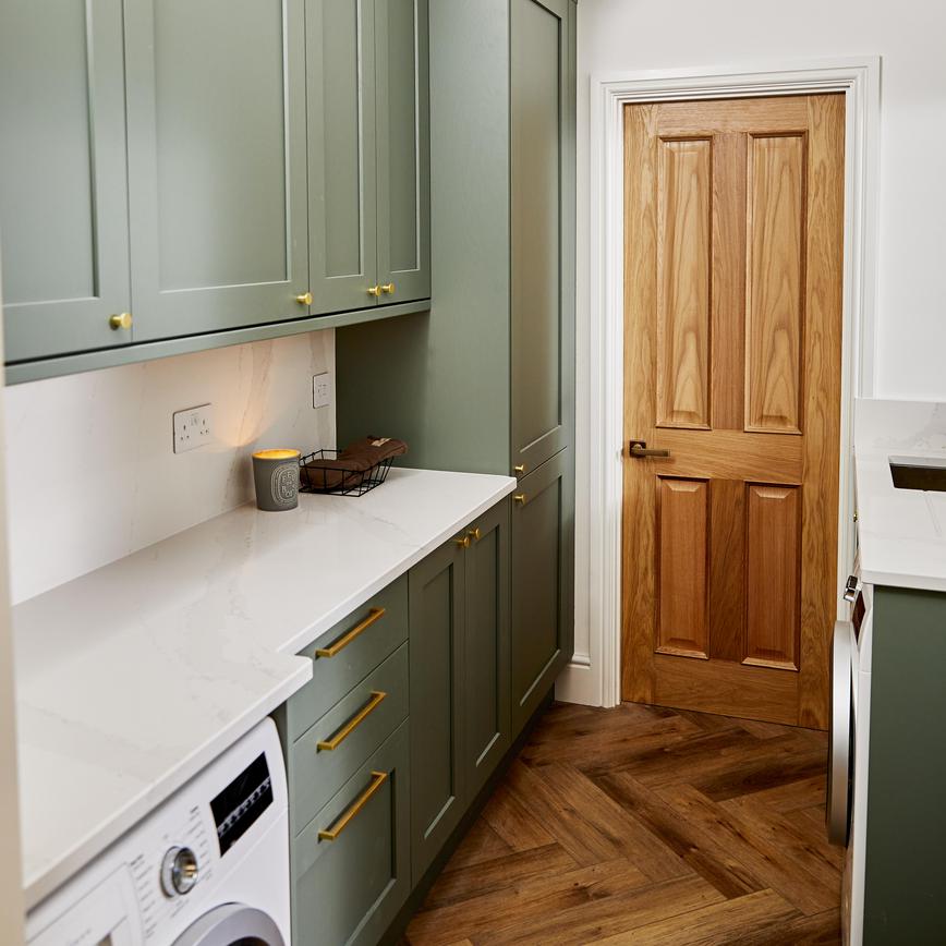 Sage green shaker utility room design with white quartz worktop, brass bar handles, a washing machine, and an oak door.
