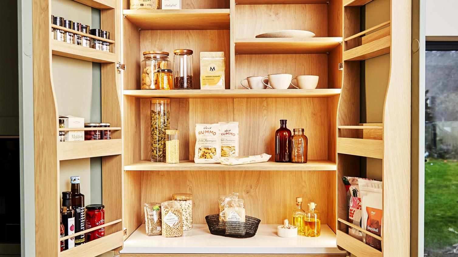Open sage green shaker larder unit with light oak shelves and door balconies showing dried food and condiments.