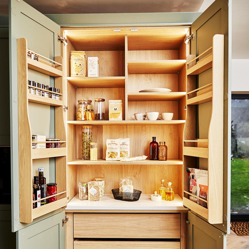 Open sage green shaker larder unit with light oak shelves and door balconies showing dried food and condiments.