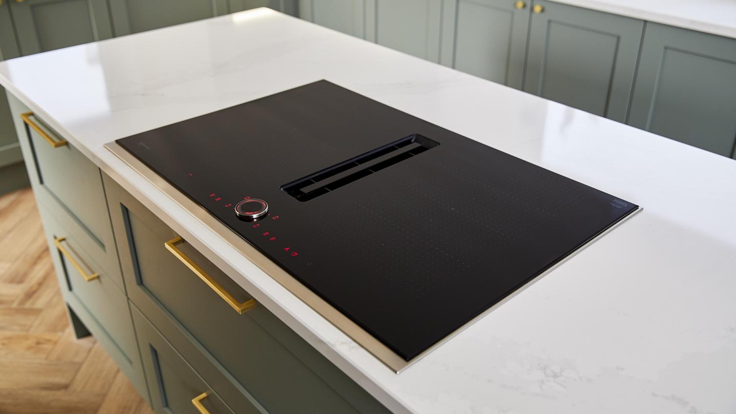 Black induction hob with a shiny surface on a reed green shaker island, with brass handles and a white quartz worktop.