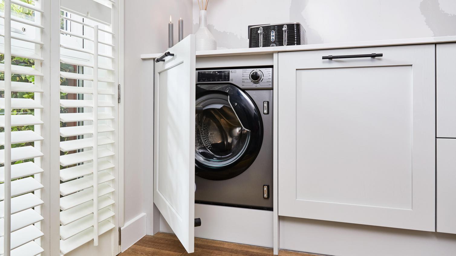 An integrated grey washing machine in a dove-grey, shaker kitchen with white, quartz worktops, black handles for wood floors.