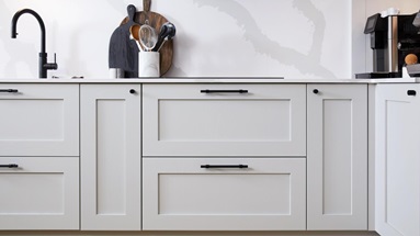 A dove-grey kitchen featuring shaker cupboard doors, black bar handles, a black tap, and white quartz worktops.