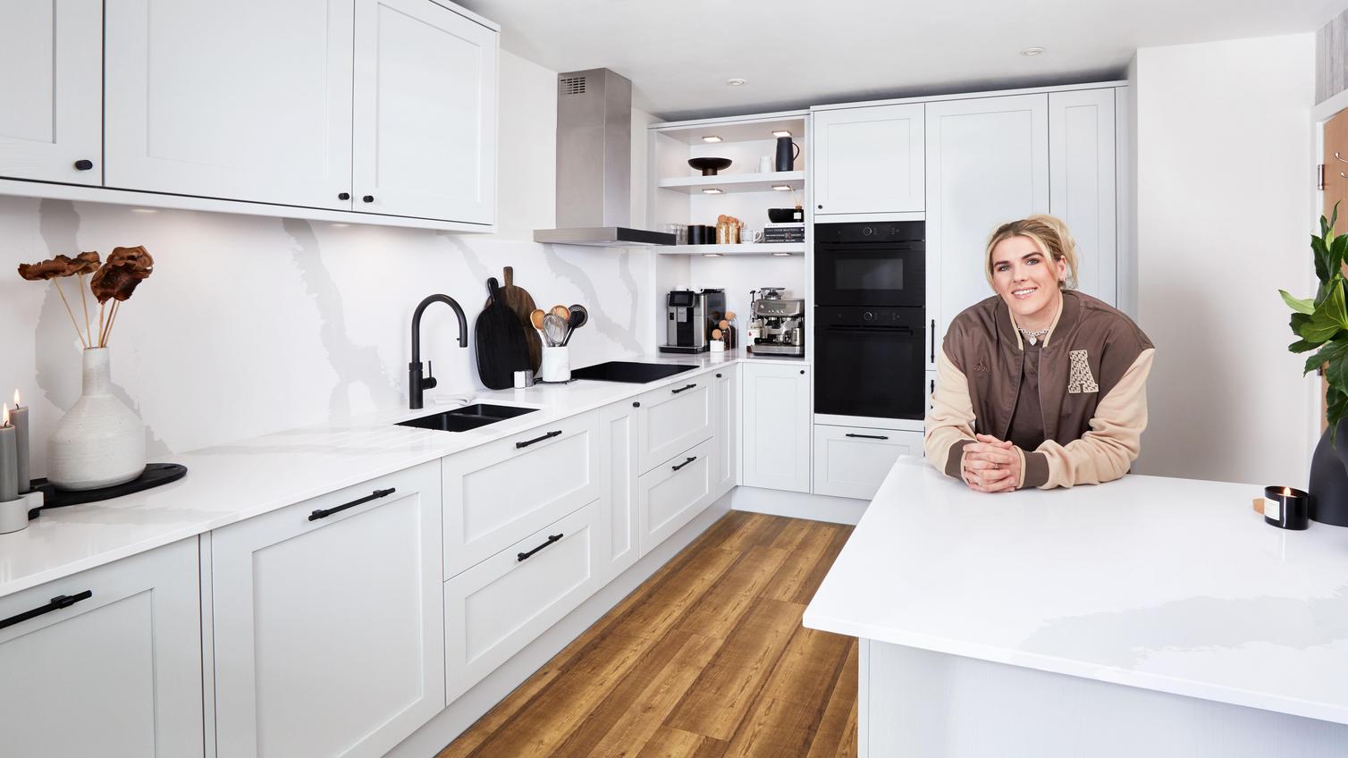 Millie Bright leaning on the breakfast bar in her new shaker kitchen, which has white worktops, black handles, and a black tap.