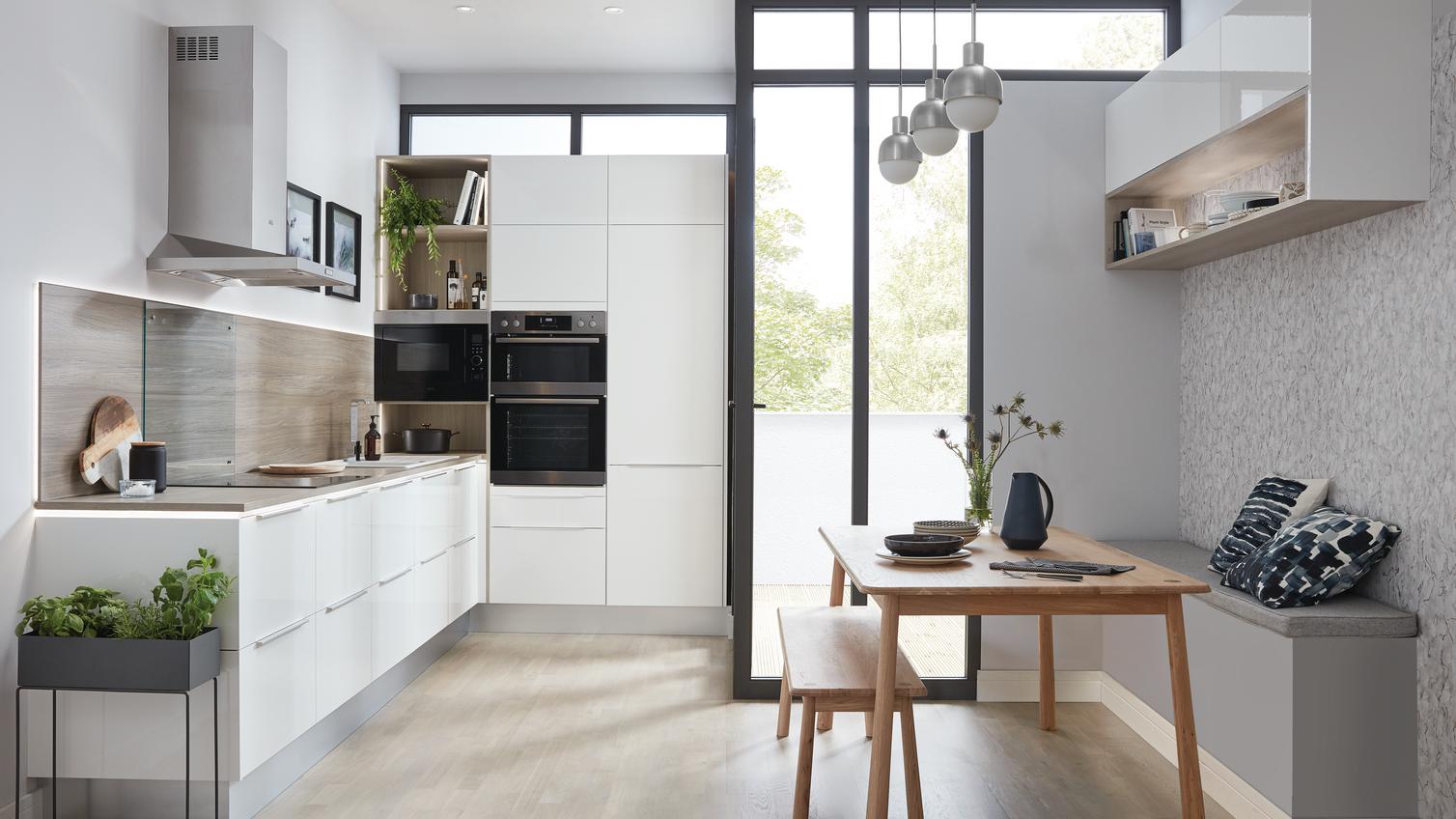 L-shape white gloss kitchen idea using slab kitchen doors. Contains a stainless-steel cooker hood and a black double oven.