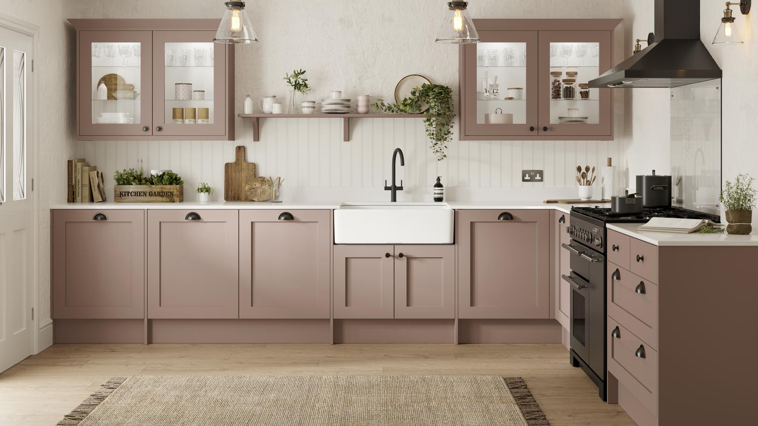 A pink, antique rose kitchen in a shaker style. It is an L-shaped layout, has light oak flooring, and black appliances.