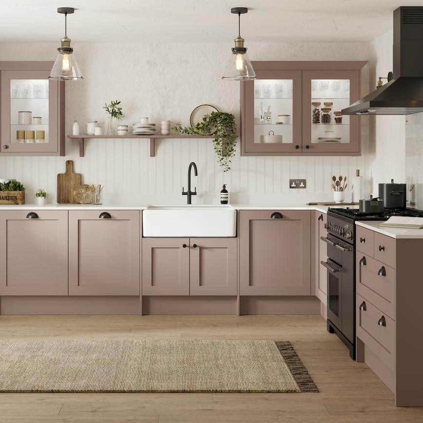 A pink, antique rose kitchen in a shaker style. It is an L-shaped layout, has light oak flooring, and black appliances.