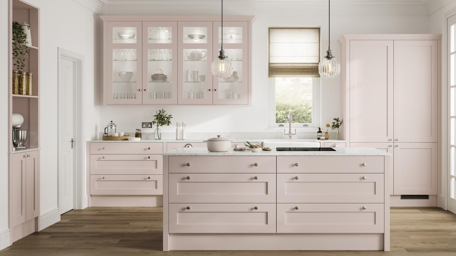 Blossom-toned, pink kitchen with shaker doors and an island layout. It has warm, oak-effect flooring and white worktops. 