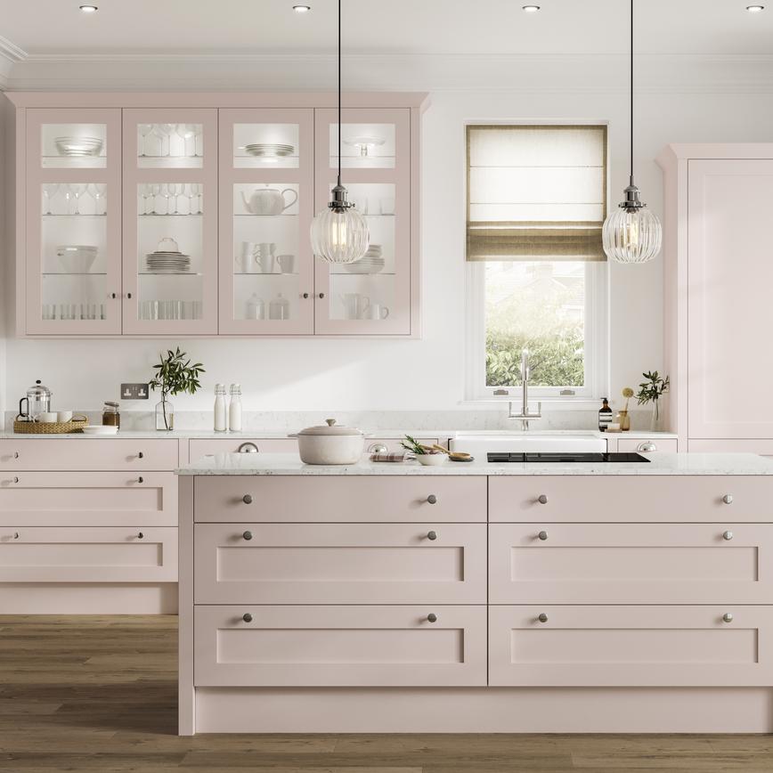 Blossom-toned, pink kitchen with shaker doors and an island layout. It has warm, oak-effect flooring and white worktops. 