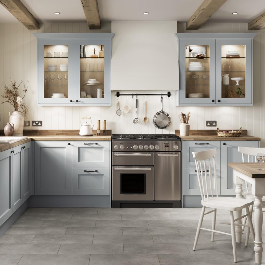 A mist blue shaker kitchen in an L-shaped layout. There are wooden worktops, two glass wall cabinets and a range cooker.