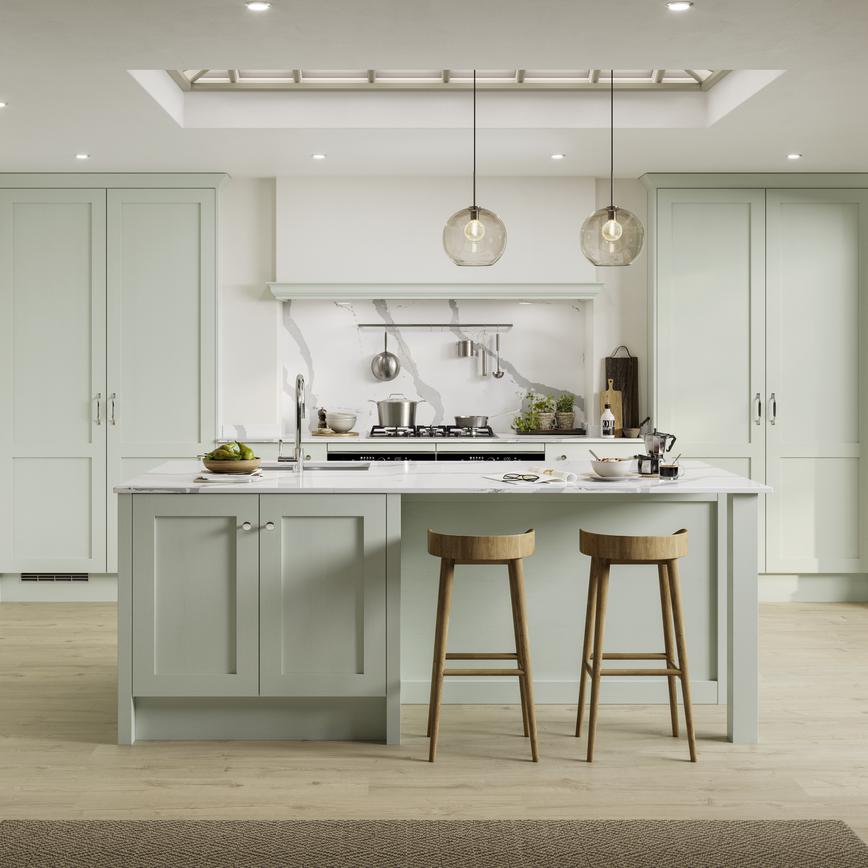 A pistachio-toned, green shaker Kitchen in an L-shaped layout with an island. It has oak-effect flooring and white worktops.