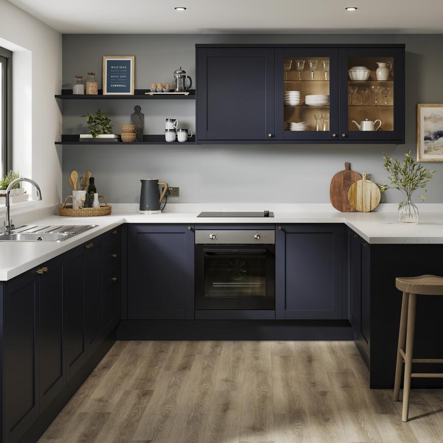 A U-shaped kitchen layout with navy, shaker doors. Includes white worktops, glass wall units, and a black, induction hob. 