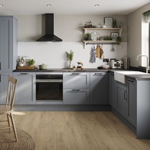 An l-shaped kitchen in a shaker style and dusk blue colour. Features oak-effect flooring and dark stone-style worktops.