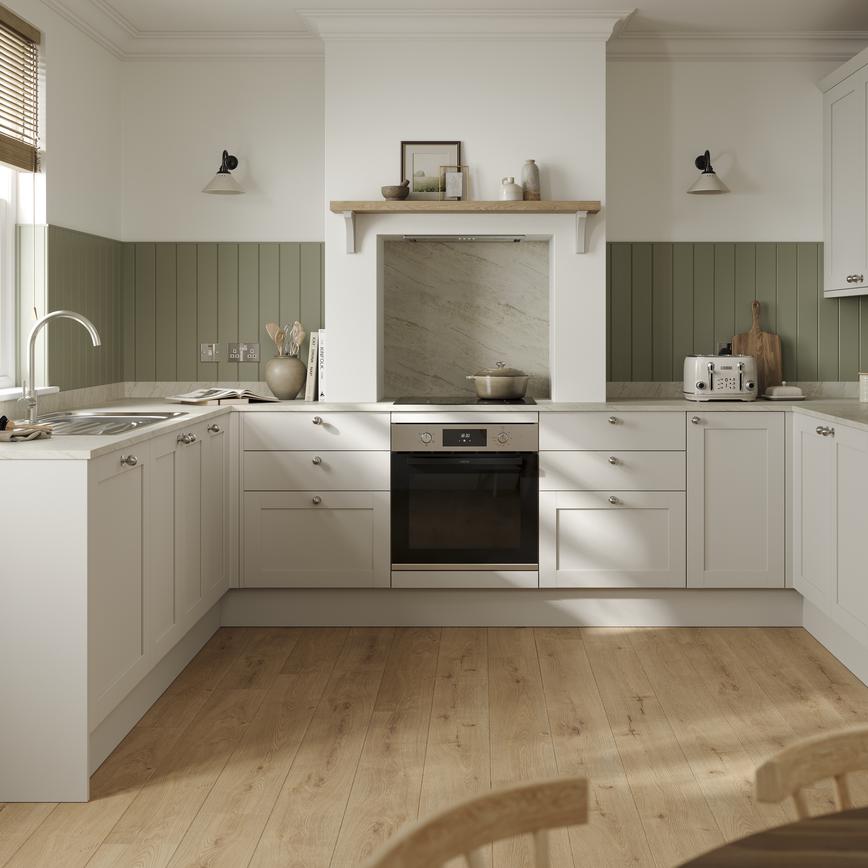 A porcelain coloured, shaker kitchen in a U-shaped layout. It has white worktops, green wall panelling, and plank flooring.