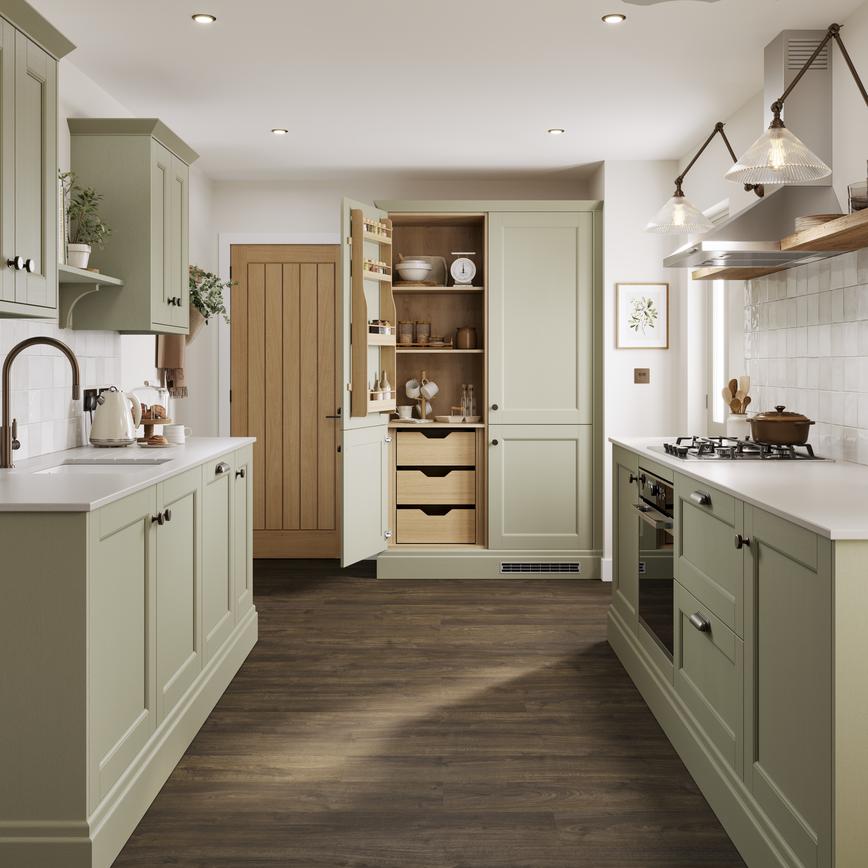 A sage green shaker kitchen with larder unit in a galley layout. It has dark oak flooring and shelving, and white worktops.