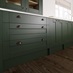 Dark-green kitchen base units, with shaker style cupboards and drawers. Showing pewter cup handles and a thin white worktop.