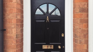 Black four-panelled door with curved glass panels at the top, brass letterbox, knocker, and knob handle, and a white frame.
