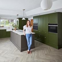 Interior influencer Kate, standing in her green and brown kitchen. It has a kitchen island as in an l-shaped layout.