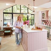 Lucy stood in her pink kitchen, leaning against the kitchen island. It has shaker doors and is styled with metallic handles.