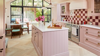 A pink shaker kitchen with an island in the centre. It is styled with metallic handles, white worktop, and oak flooring.