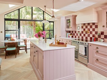 A pink shaker kitchen with an island in the centre. It is styled with metallic handles, white worktop, and oak flooring.