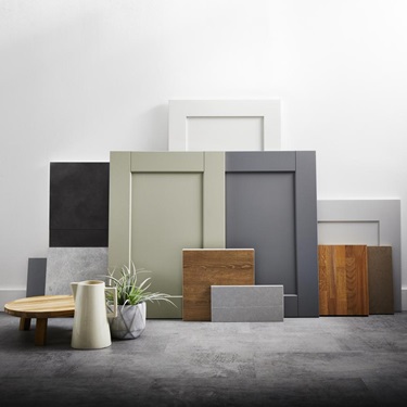 Shaker kitchen doors and boards in natural hues, like green, oak, and white on a grey floor. Propped up against a white wall.