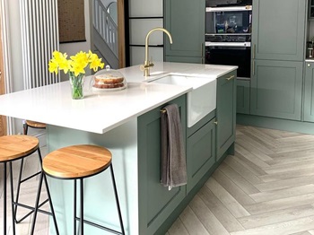 Green shaker kitchen island idea with a Belfast sink, brass tap, white worktops, and chevron flooring.