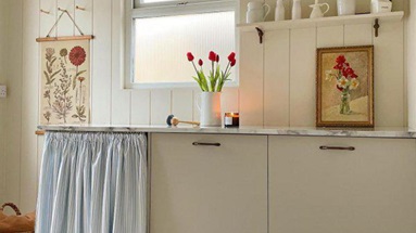 A slab kitchen in a subtle sandstone colour, with a blue curtain, red flowers and cream wall panelling