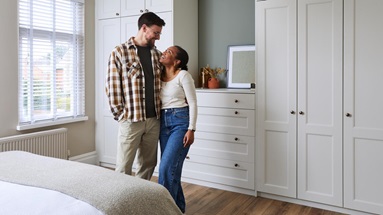 Jade and James standing in their new Bridgemere bedroom. It has oak vinyl flooring, tall wardrobes and matching drawer units