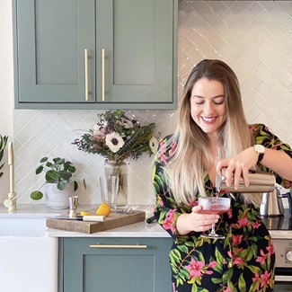 Poppy stood in front of a teal shaker kitchen, wearing a floral dress, looking down, and pouring a cocktail into a glass.